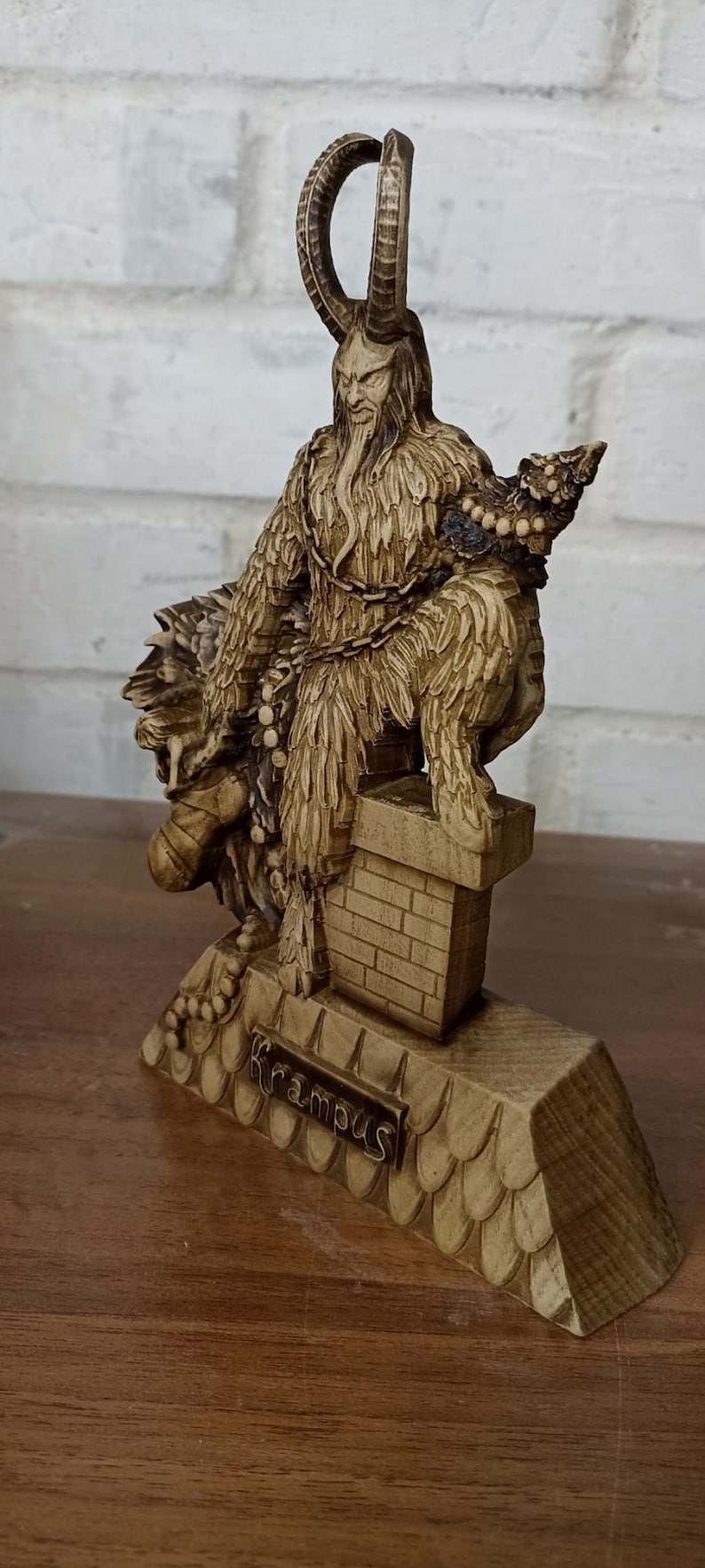 May include: A detailed, tan-colored Krampus figurine. The figure has large horns, a long beard, and is chained. The base is inscribed with the word "Krampus". The figurine is on a wooden surface.