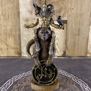 Puede incluir: Estatua detallada que presenta una figura con múltiples cabezas de animales sobre un oso, sosteniendo un cuervo. La estatua es marrón y dorada, con una base circular con símbolos. La estatua está sobre una base de madera.