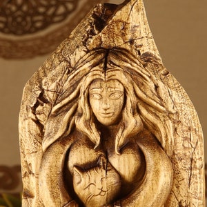 May include: A wooden carving of a woman with long hair holding a cat. The woman is carved in a detailed style with a flowing robe and a serene expression. The cat is nestled in her arms and is also carved with intricate details.