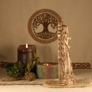 May include: A wooden wall hanging featuring a carved depiction of a goddess figure with flowing hair and a long dress. The figure is surrounded by intricate carvings of leaves and vines. The wall hanging is set against a backdrop of a brown fabric with a woven tree of life design.