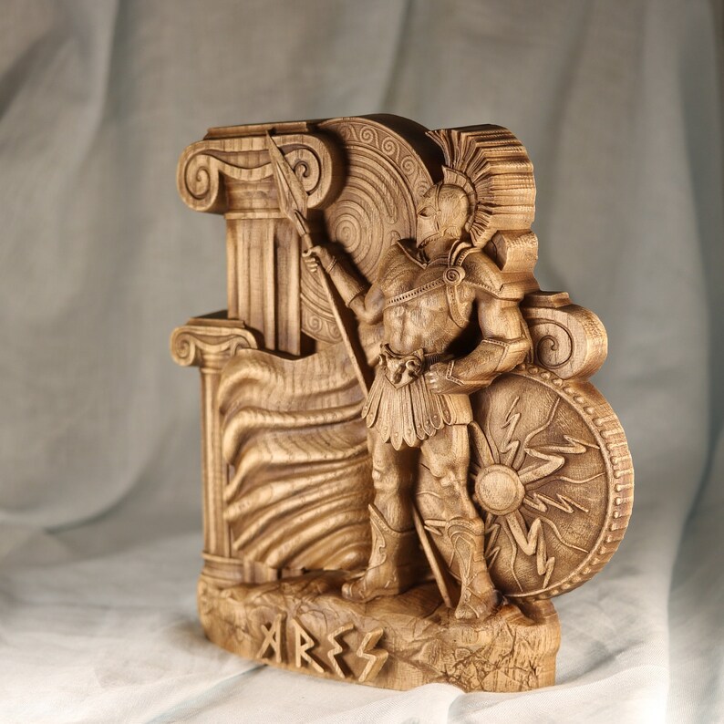May include: A wooden carving of the Greek god Ares, the god of war. He is depicted in a warrior pose, holding a spear and shield. The carving is detailed and intricate, with a flowing cape and a helmet with a plume. The carving is labeled "Ares" at the bottom.