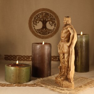 May include: A wooden carving of a Viking warrior standing on a wooden base. The carving is detailed and realistic, with a beard, a cloak, and a sword. The carving is surrounded by three candles, two brown and one green.
