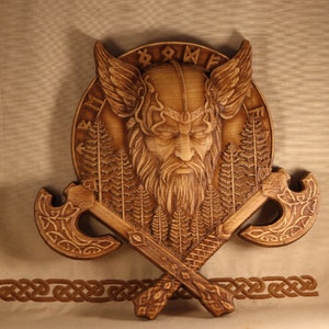 May include: A wooden wall hanging featuring a carved Viking warrior head with a beard and helmet, surrounded by a circle of runes and trees. Two crossed axes are attached to the bottom of the carving.