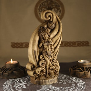 Op de afbeelding: Een houten sculptuur van een vrouw die in een wervelende, wolkachtige formatie staat. De vrouw draagt een hoofdtooi en een decoratieve riem. Het beeld is gebeeldhouwd met ingewikkelde details en heeft een warme bruine kleur.