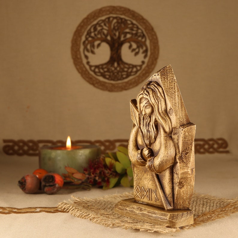 May include: A wooden carving of a Norse god, possibly Odin, with a long beard and holding a staff. The carving is on a wooden base with runes inscribed on it. The carving is in front of a candle and a wooden background.