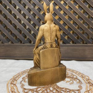May include: Wooden sculpture of a seated figure with rabbit ears. The figure is carved in a realistic style and has a detailed, textured surface. The sculpture is painted in a gold color.