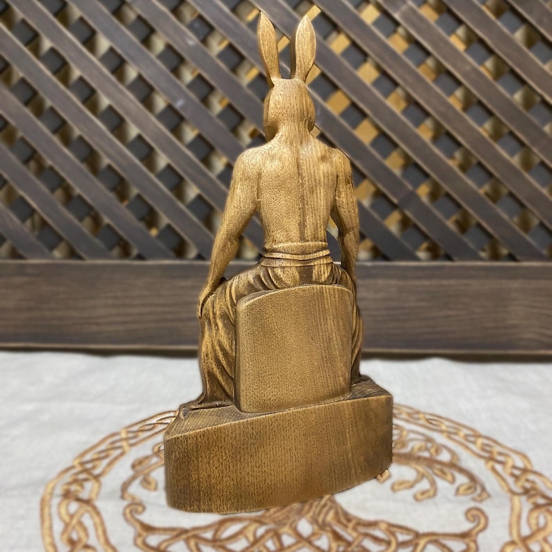 May include: Wooden sculpture of a seated figure with rabbit ears. The figure is carved in a realistic style and has a detailed, textured surface. The sculpture is painted in a gold color.