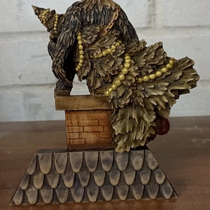 May include: A detailed figurine of a horned creature with a fur-like body, perched atop a brick chimney. The figure is adorned with gold-colored beads and sits on a roof-like base, showcasing intricate carvings.