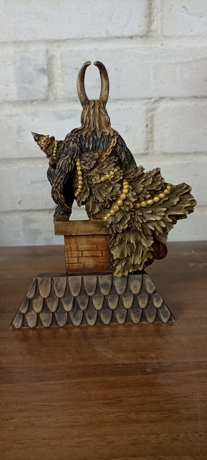 May include: A detailed figurine of a horned creature with a fur-like body, perched atop a brick chimney. The figure is adorned with gold-colored beads and sits on a roof-like base, showcasing intricate carvings.