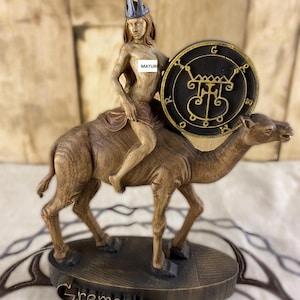 Könnte beinhalten: Detaillierte Holzskulptur einer Figur, die ein Kamel reitet. Die Figur trägt eine Krone und hält einen Schild mit komplizierten Symbolen. Die Basis der Skulptur ist mit dem Namen "Gremory" beschriftet. Das Wort "MATURE" ist ebenfalls sichtbar.