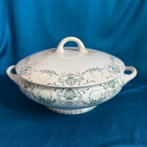 May include: A white ceramic lidded serving dish with a green floral and shell pattern. The dish has two handles and a loop handle on the lid. The dish sits on a small pedestal base.