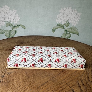 May include: A rectangular box with a white background and a floral pattern in red, green, and grey. The box has a light brown border.