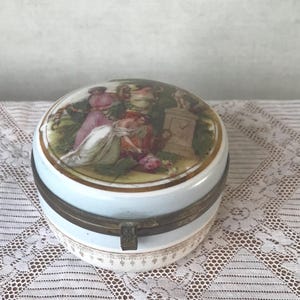 May include: A round porcelain trinket box with a hinged lid. The lid features a painted scene of three women in a garden setting. The box is decorated with gold trim and a blue band around the middle.
