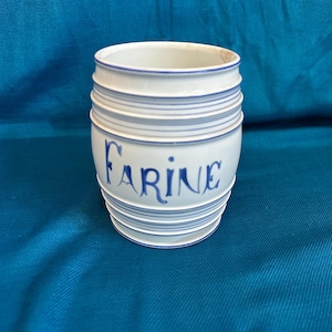 May include: A white ceramic jar with blue horizontal stripes and the word "Farine" in blue script. The jar has a barrel shape and is likely used for storing flour. The background is a solid teal colour.