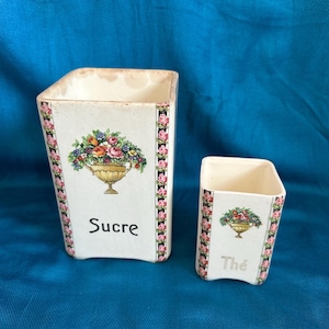 May include: Two white ceramic containers with floral designs and the words "Sucre" and "Thé". The containers have a rectangular shape and are decorated with a border of pink roses. The larger container has a floral arrangement in a gold-coloured vase.