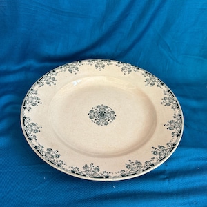 May include: A round ceramic plate with a cream-coloured base featuring a dark green floral pattern along the rim and a matching circular floral design centred in the middle of the plate. The plate has a smooth, glossy finish and a wide, flat border.