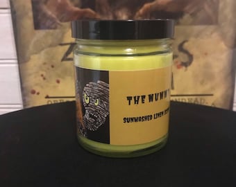 May include: A yellow candle in a glass jar with a black lid. The label on the jar features a cartoon mummy with the text "THE MUMMY" and "SUNWASHED LINEN SCENT".