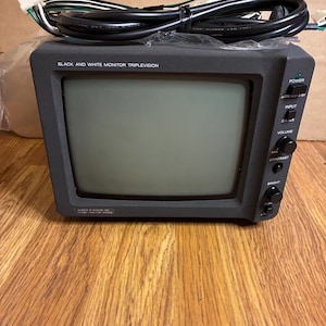 May include: A black and white Trilevision monitor with a square screen. The monitor has several control knobs and buttons on the right side, including power, input, volume, contrast, and brightness. A black cable is coiled on top.