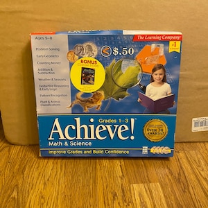 Puede incluir: Una caja azul y amarilla para el software educativo Achieve! Math & Science para los grados 1-3. La caja presenta a una niña mirando a un lagarto a través de una lupa. La caja también incluye el texto "Ages 5-8" y "Bonus CD-ROM Inside!" y "Over 30 Awards!"