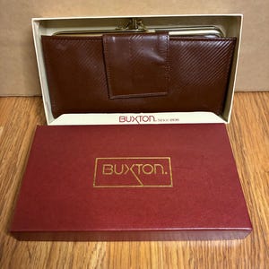 May include: A brown leather wallet with a clasp closure, presented in an open white box. The wallet has a textured surface and is displayed on a wooden surface. The box is red with the gold text "BUXTON."