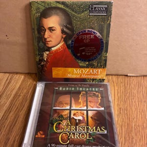 Puede incluir: Se muestran dos CD. El CD superior se titula "MOZART Musical Masterpieces" y presenta un retrato de Mozart. El CD inferior es "A Christmas Carol" de Dickens, una producción dramática completa de 90 minutos.