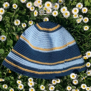 Puede incluir: Un sombrero de ganchillo en tonos azules y dorados, sobre un lecho de hierba verde y margaritas blancas. El sombrero presenta un diseño a rayas con una base azul oscuro, un centro azul claro y detalles dorados. Un accesorio artesanal único.