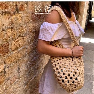 Puede incluir: Un bolso tote de crochet beige de tejido abierto con una correa larga. El bolso lo sostiene una persona que lleva un vestido blanco. El bolso tiene forma cilíndrica y está hecho de hilo de color natural. El texto "狐狸" es visible.