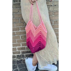 Puede incluir: Un bolso tote de ganchillo rosa y melocotón con un patrón de chevrón. El bolso hace la transición de melocotón claro en la parte superior a un magenta intenso en la parte inferior. El bolso tiene correas largas y lo sostiene una persona.