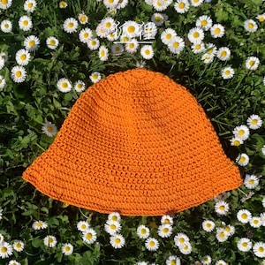 Puede incluir: Un sombrero de cubo naranja tejido a ganchillo, sobre un lecho de hierba verde y margaritas blancas. El sombrero tiene un ala ancha y un aspecto texturizado. Este sombrero para el sol es de color liso.