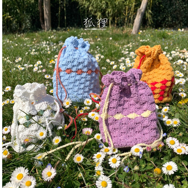 Puede incluir: Cuatro bolsas de crochet hechas a mano con cord&oacute;n en blanco, azul claro, morado y amarillo. Cada bolsa presenta un patr&oacute;n de puntada texturizada y un acento de color contrastante cerca de la parte superior. Las bolsas est&aacute;n en un campo de margaritas.