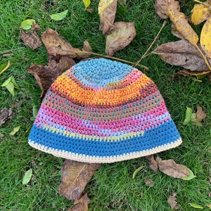 Puede incluir: Un gorro de ganchillo hecho a mano con un diseño de rayas coloridas. El gorro presenta bandas azules, rosas, amarillas y naranjas, con un borde color crema. El gorro está sobre hierba verde y hojas caídas.