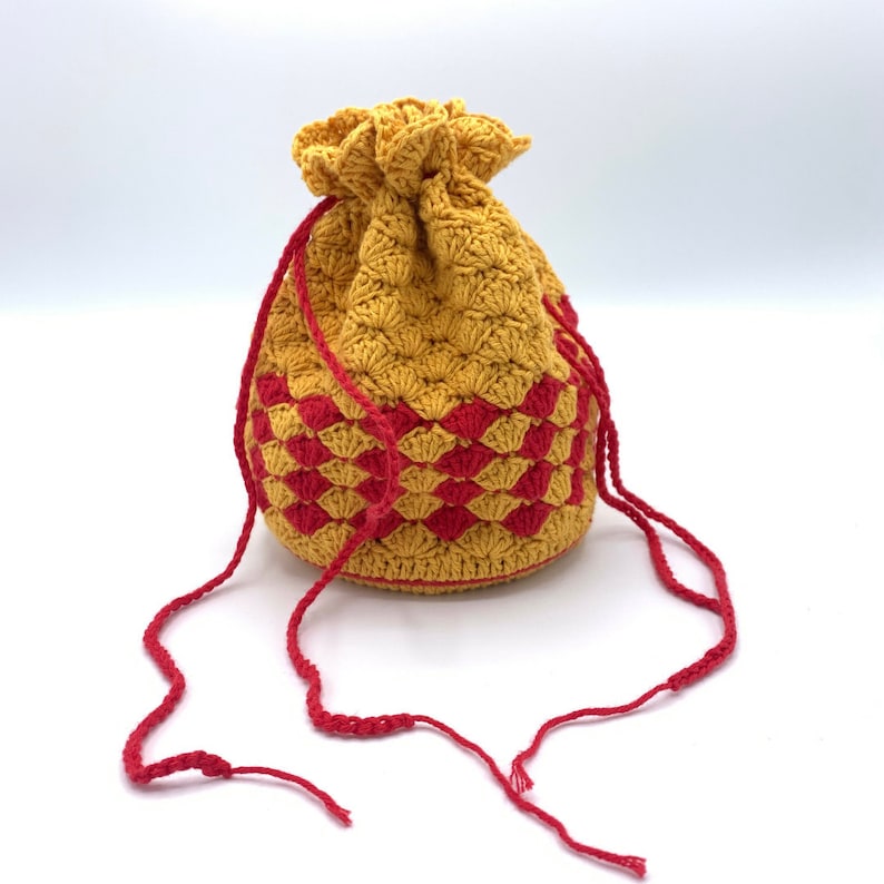 Puede incluir: Un bolso de ganchillo hecho a mano con cord&oacute;n en una combinaci&oacute;n de colores amarillo dorado y rojo. El bolso presenta un patr&oacute;n texturizado y un cierre de cord&oacute;n rojo. El bolso mide aproximadamente 15 cm de alto y 13 cm de ancho.