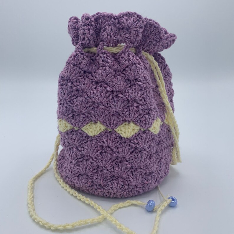 Puede incluir: Un bolso con cord&oacute;n hecho a mano, de ganchillo morado, con una banda de acento color crema. El bolso tiene un cord&oacute;n a juego y un peque&ntilde;o detalle de cuentas azules. El bolso est&aacute; dise&ntilde;ado con un patr&oacute;n de puntada texturizada.