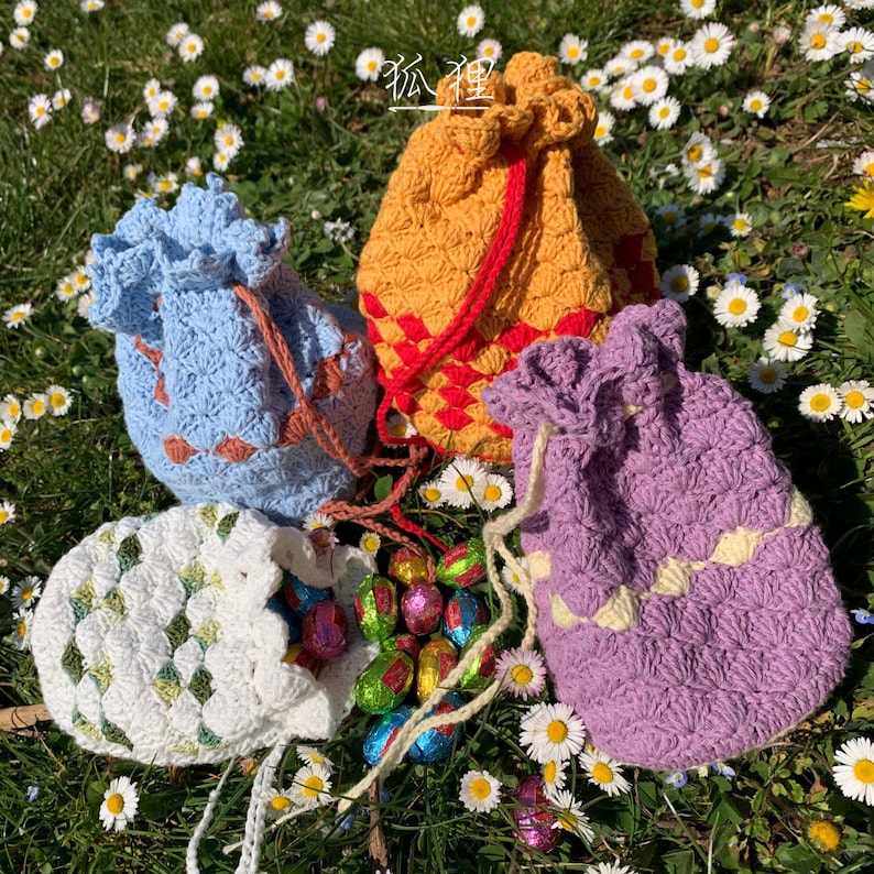 Puede incluir: Cuatro bolsas de cord&oacute;n de ganchillo en varios colores: azul claro, naranja, morado y blanco. Las bolsas est&aacute;n llenas de huevos de chocolate y descansan sobre hierba y margaritas. Cada bolsa tiene un patr&oacute;n de puntada &uacute;nico y un cord&oacute;n.