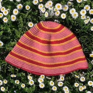 Puede incluir: Sombrero de ganchillo con rayas rosas, rojas y amarillas. El sombrero está sobre un lecho de hierba verde y margaritas blancas.