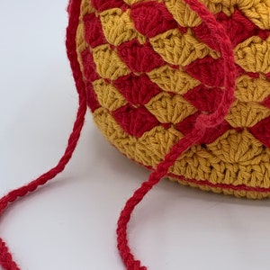 Puede incluir: Un bolso de ganchillo con forma redondeada, con un patr&oacute;n repetido de formas de diamante doradas y rojas alternas. El bolso tiene una correa roja a juego. La textura del ganchillo es visible y el bolso parece hecho a mano.