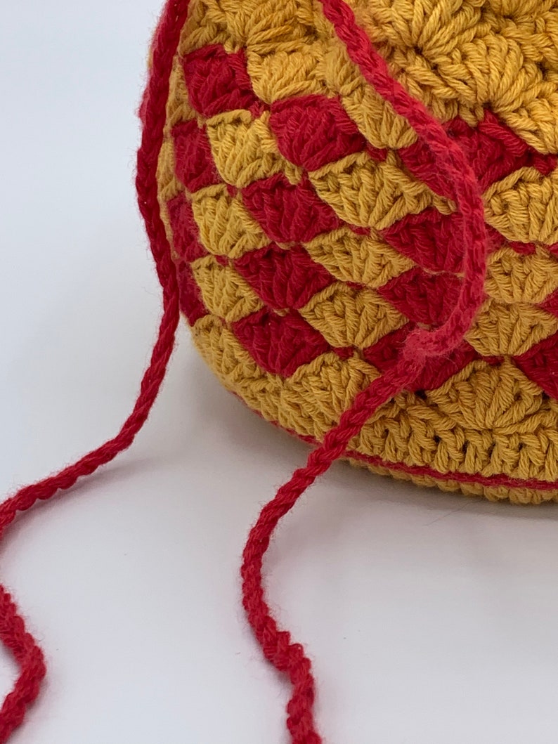 Puede incluir: Un bolso de ganchillo con forma redondeada, con un patr&oacute;n repetido de formas de diamante doradas y rojas alternas. El bolso tiene una correa roja a juego. La textura del ganchillo es visible y el bolso parece hecho a mano.