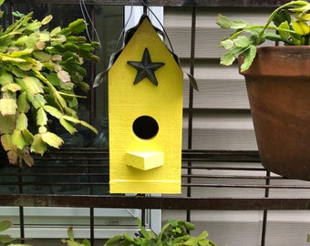 May include: A yellow birdhouse with a black star on the front and a small opening for birds to enter. The birdhouse is hanging from a metal rack.