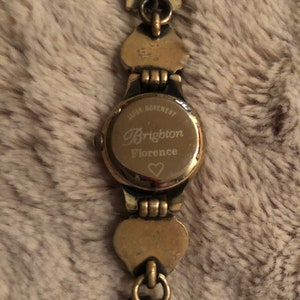 May include: Gold-tone watch with a heart-shaped design on the band. The watch face has the text "Japan Movement Brighton Florence" and a small heart symbol.