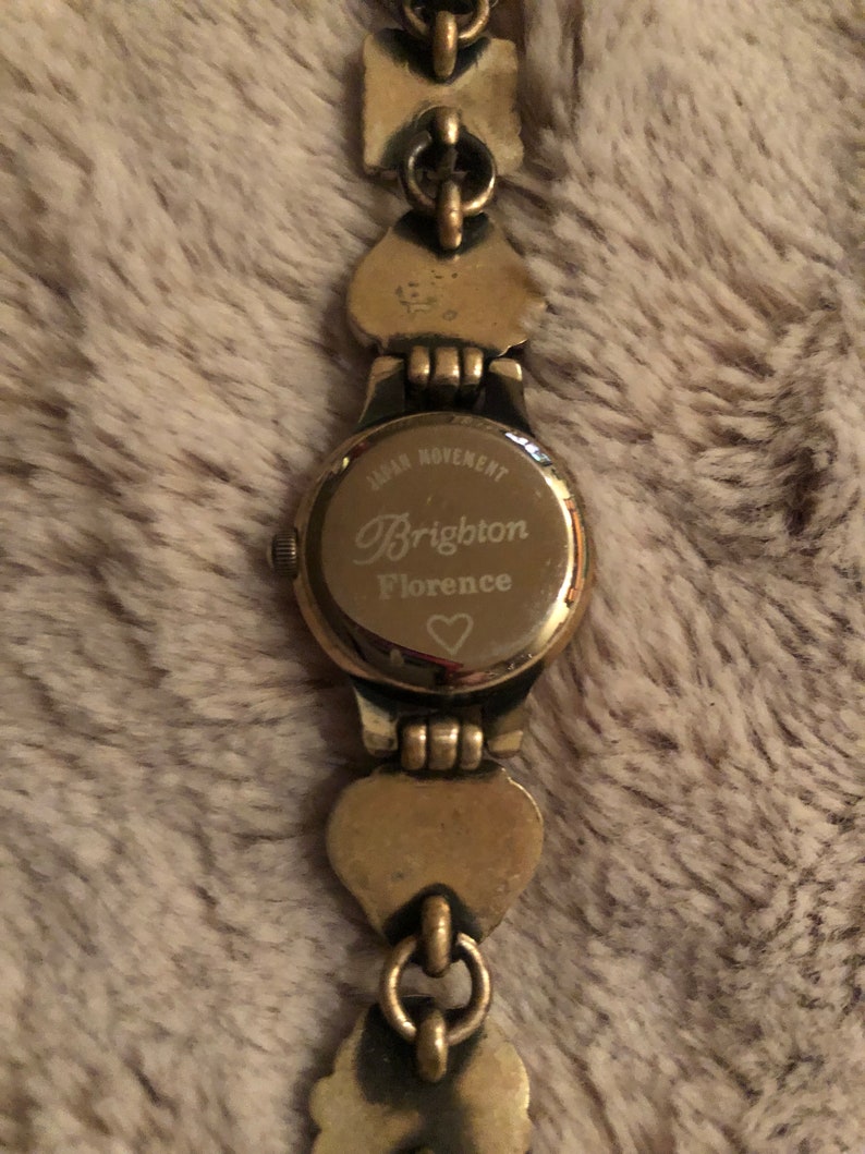 May include: Gold-tone watch with a heart-shaped design on the band. The watch face has the text "Japan Movement Brighton Florence" and a small heart symbol.