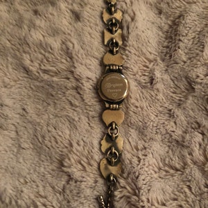 May include: Gold-tone watch with a heart-shaped design. The watch face has the text "Brighton Florence" with a small heart symbol.