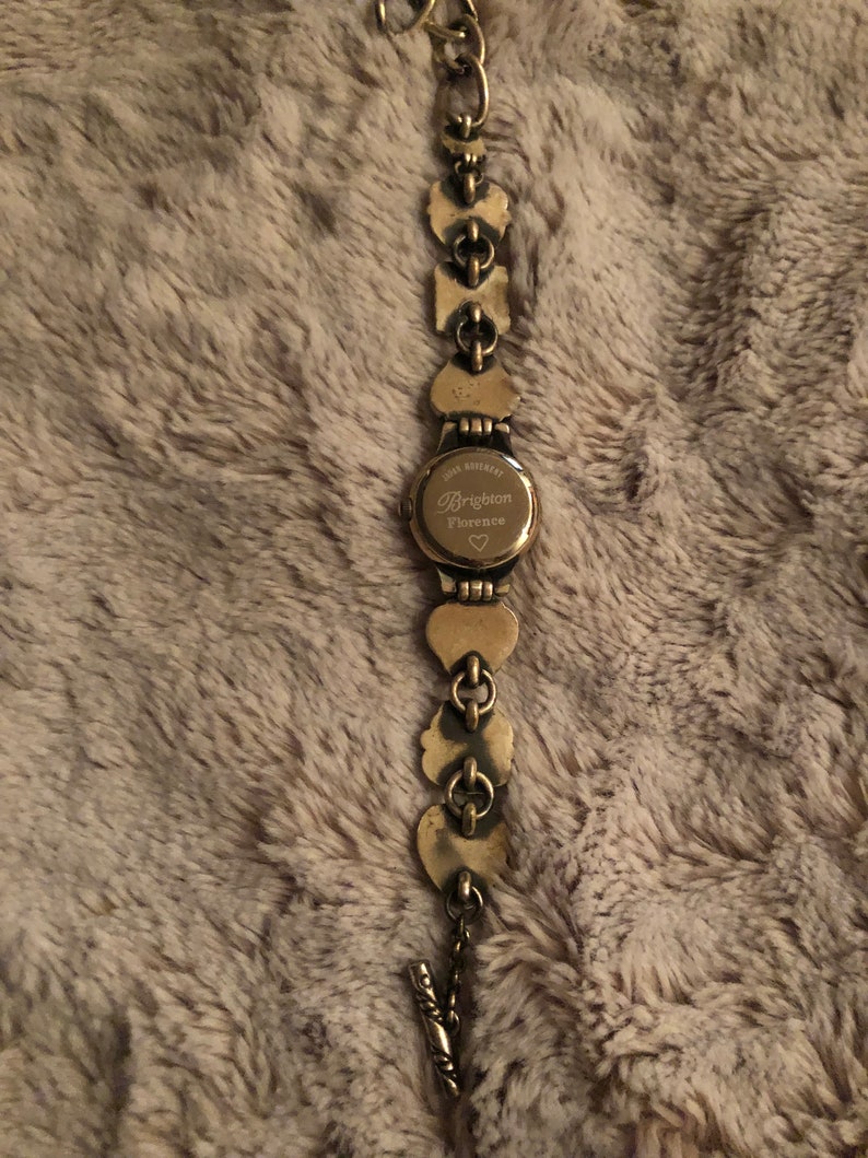 May include: Gold-tone watch with a heart-shaped design. The watch face has the text "Brighton Florence" with a small heart symbol.