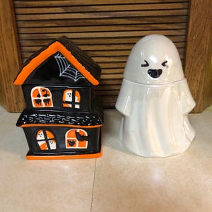 May include: Two Halloween themed ceramic containers. One is a black and orange haunted house with a spider web on the roof. The other is a white ghost with a smiling face.