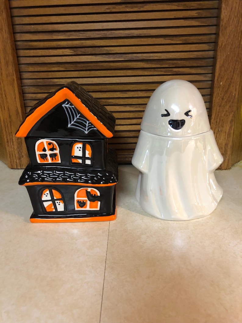 May include: Two Halloween themed ceramic containers. One is a black and orange haunted house with a spider web on the roof. The other is a white ghost with a smiling face.