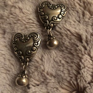 May include: A pair of silver heart-shaped dangle earrings with a detailed swirl design. The earrings have a small ball charm hanging from the bottom of each heart.