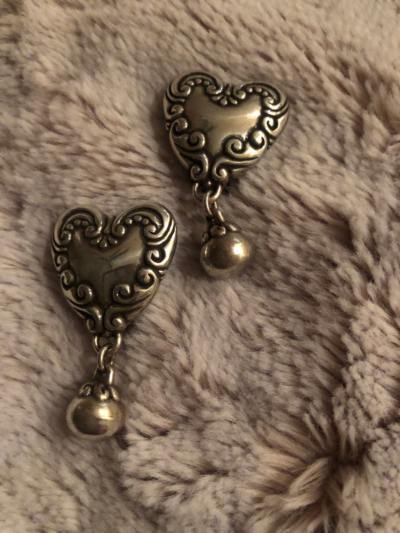 May include: A pair of silver heart-shaped dangle earrings with a detailed swirl design. The earrings have a small ball charm hanging from the bottom of each heart.