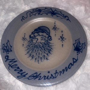 May include: A round ceramic plate with a speckled light gray finish. The center features a hand-painted blue illustration of a Santa Claus face surrounded by small star accents. The rim displays blue holly leaves and the text Merry Christmas.