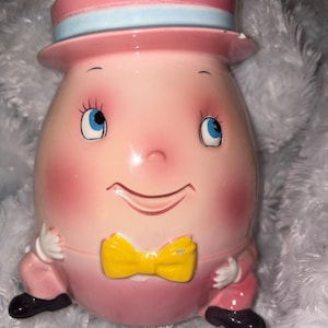May include: A ceramic Humpty Dumpty figurine featuring a pink egg-shaped body, a pink top hat with a light blue band, a yellow bow tie, and black shoes. The face has blue eyes, long eyelashes, and rosy cheeks, with white hands resting on the sides.