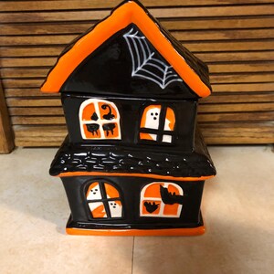May include: A black and orange ceramic haunted house with a white spider web on the roof. The house has four windows with ghosts and bats inside.