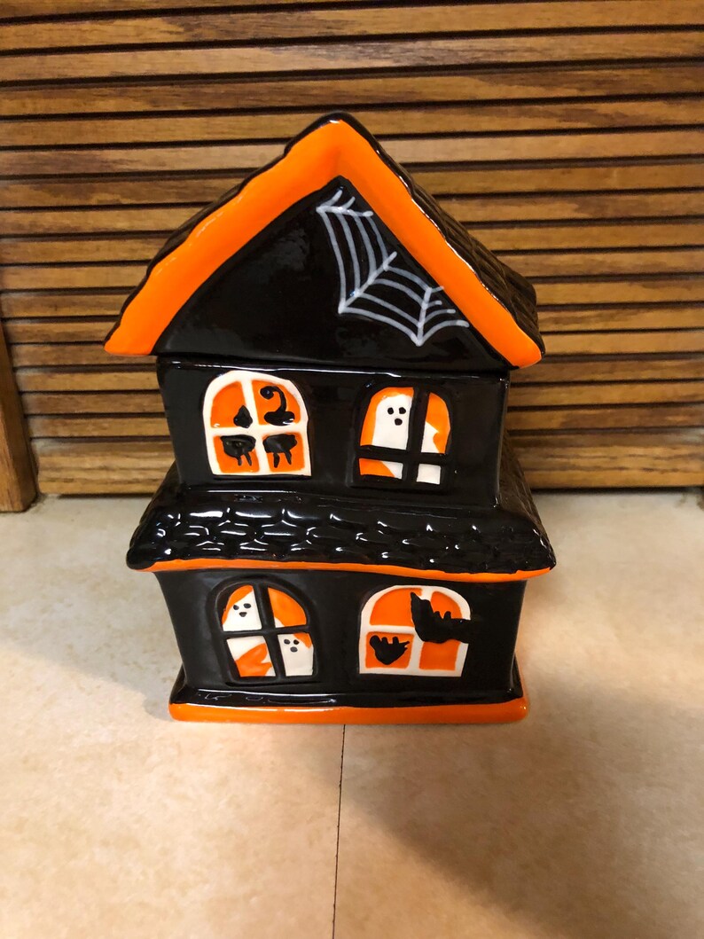 May include: A black and orange ceramic haunted house with a white spider web on the roof. The house has four windows with ghosts and bats inside.